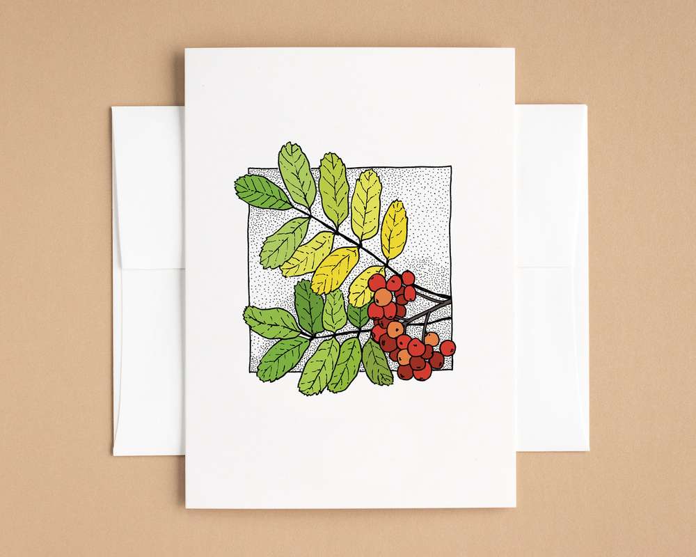 A white vertical greeting card, depicting two branches of green and yellow leaves and a spray of red berries. The card sits on top of a white envelope, and both sit on a brown backdrop.